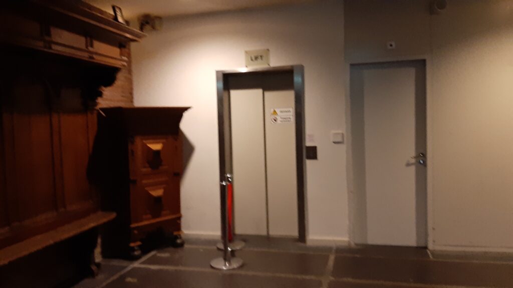 The lift door is narrow and barred on the far side by a velvet rope that protects an old piece of furniture.
