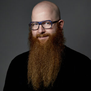 A man in a black shirt, shown from the chest up. He is bald, wears big glasses, and has a substantial red beard.